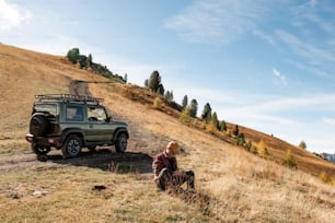 a man sitting on the side of a dirt road next to a green jeep