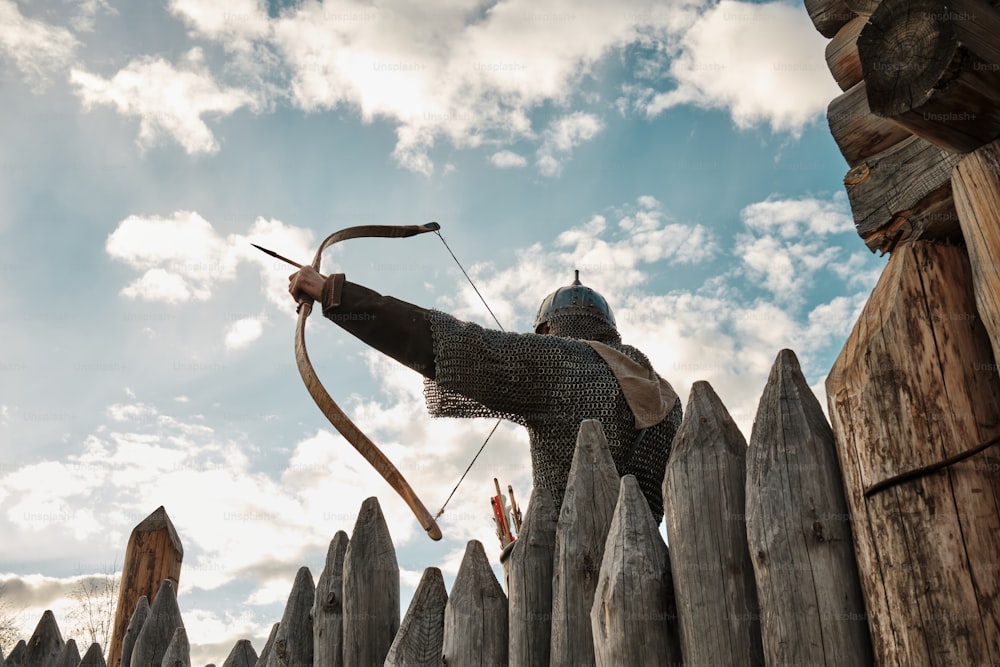 Un hombre con arco y flecha parado frente a una valla de madera
