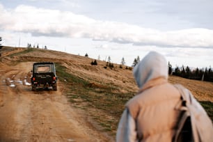 a person in a hooded jacket is looking at a jeep on a dirt road