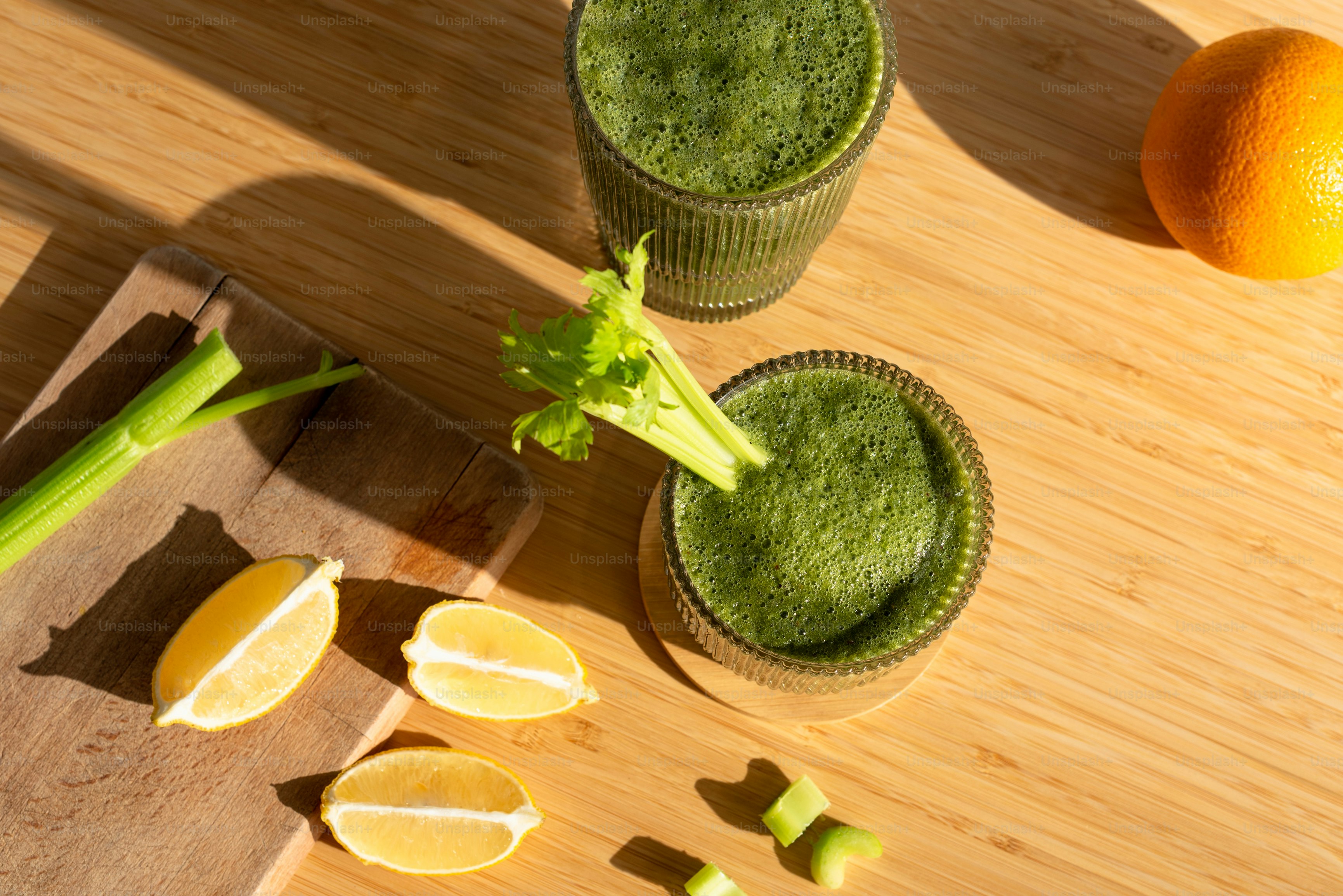 two glasses of green smoothie next to an orange and celery