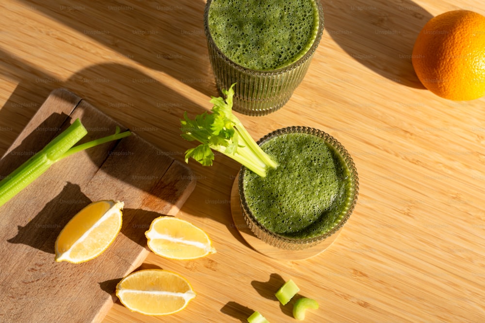 two glasses of green smoothie next to an orange and celery