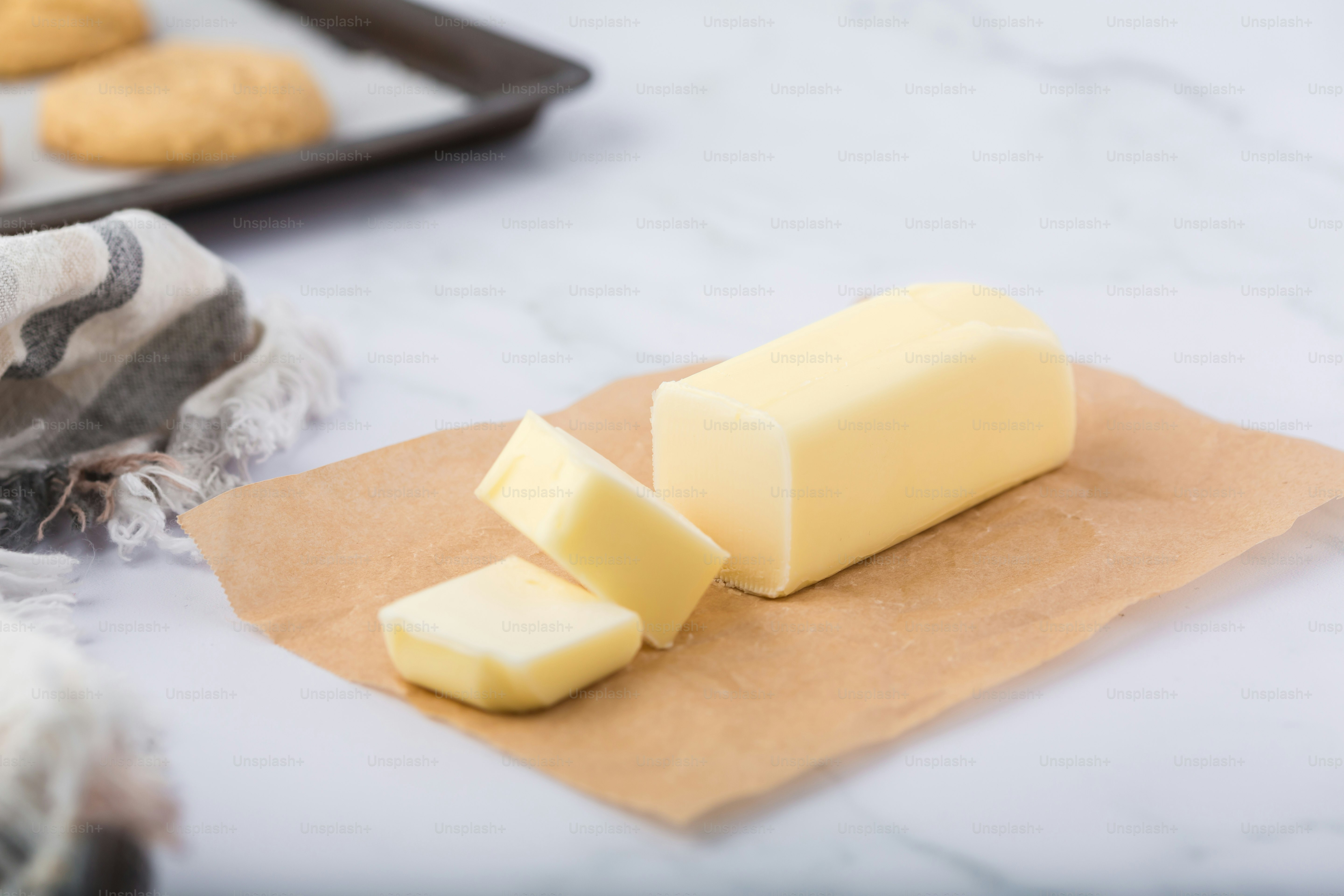 A sliced stick of butter sits on parchment paper with a linen napkin nearby and a baking sheet with cookies in the background.