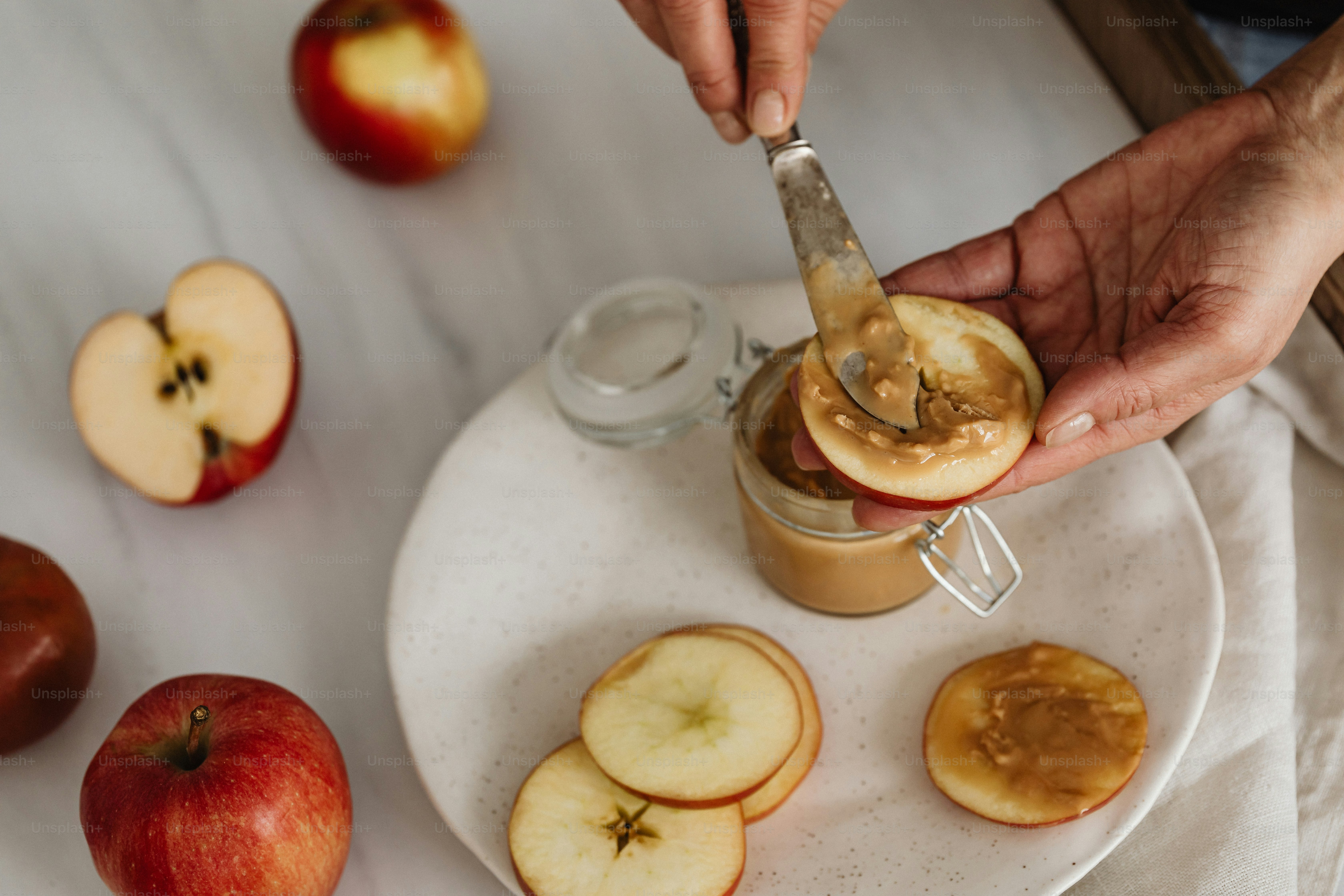 a person holding a knife over a jar of peanut butter and apples