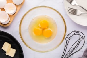バターの泡立て器の隣のボウルに3つの卵があります