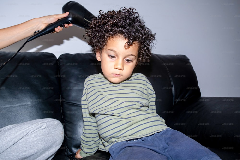 a little boy sitting on a couch with a hair dryer on his head