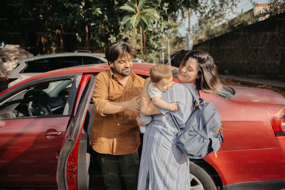 a man and woman holding a baby in front of a red car