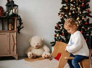 a little girl sitting on a rocking horse next to a christmas tree