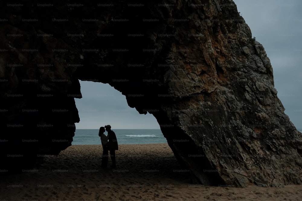 a couple standing under a large rock on a beach