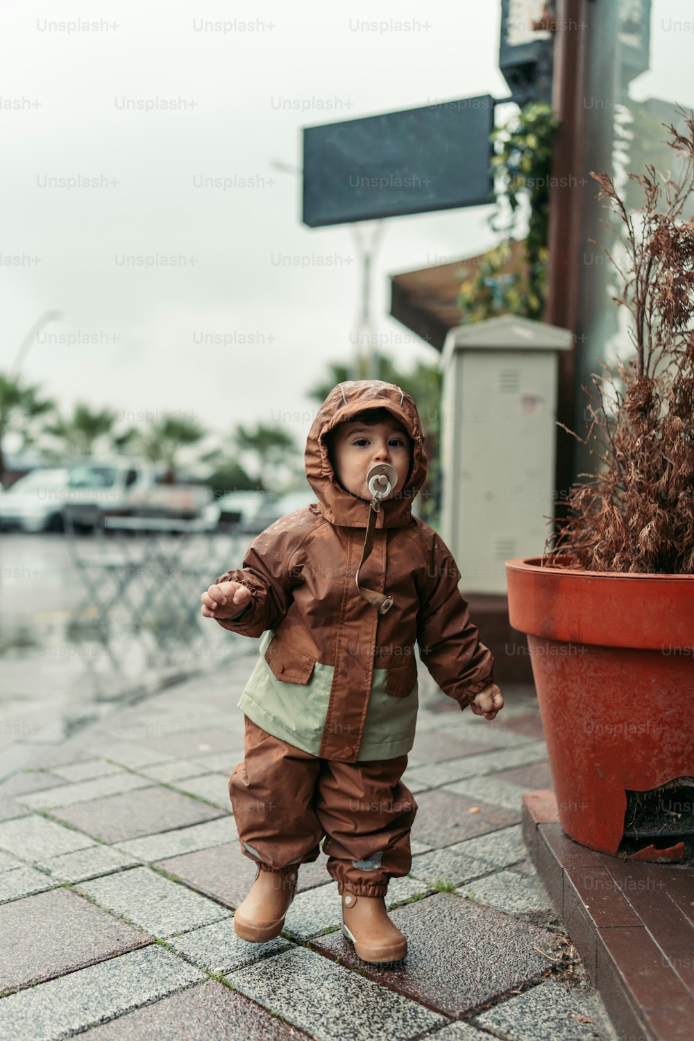 a toddler in a brown snowsuit standing on a sidewalk