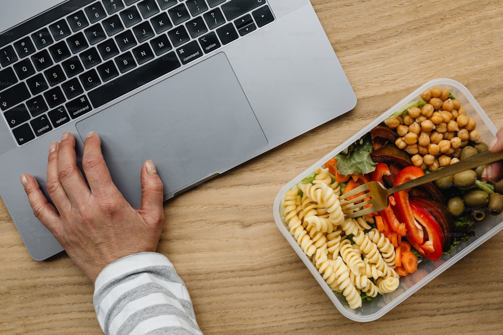 a person is using a laptop and eating pasta salad