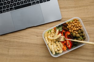 a plastic container filled with pasta salad next to a laptop