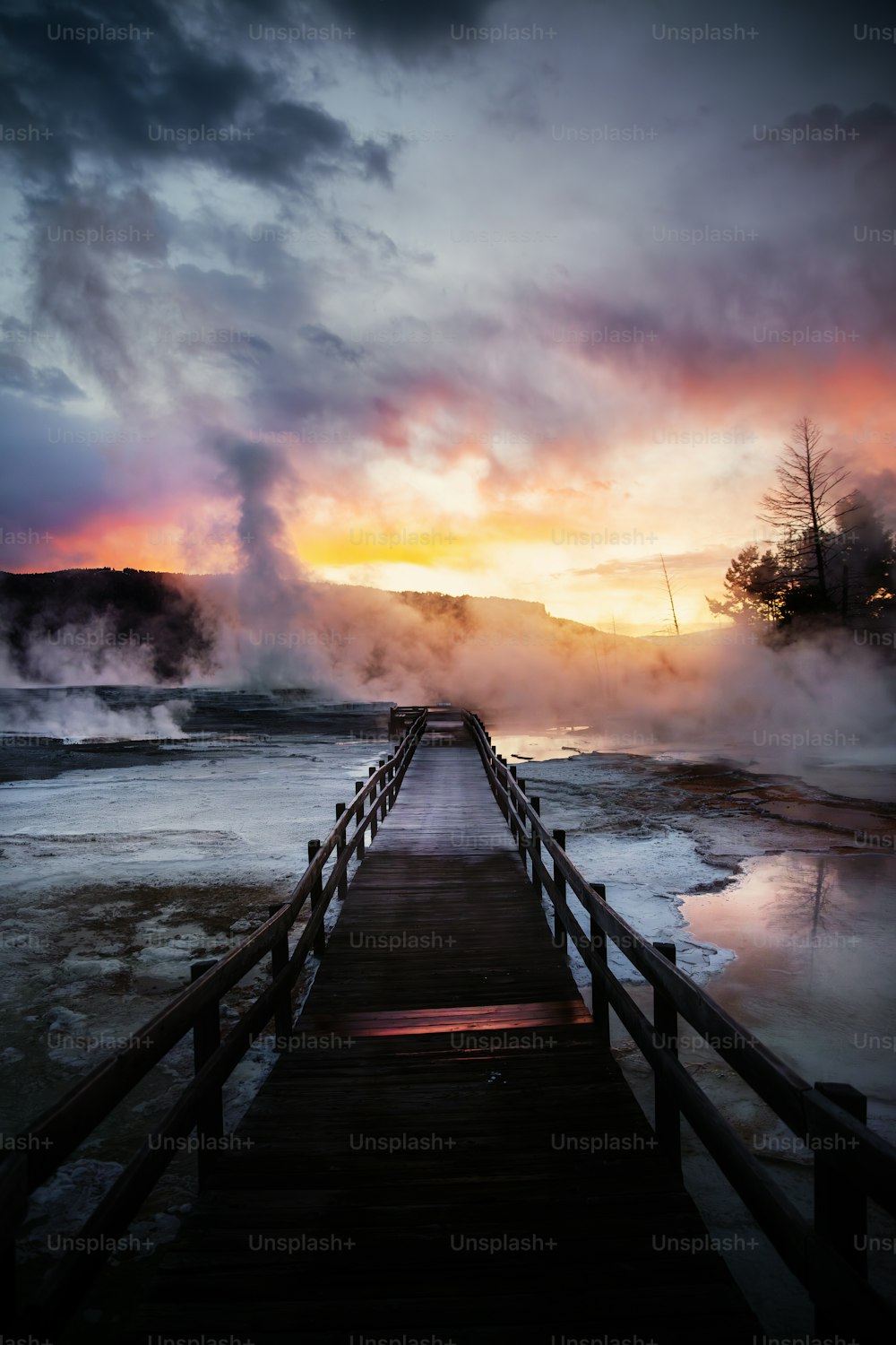 a boardwalk leading to a steaming geyser at sunset