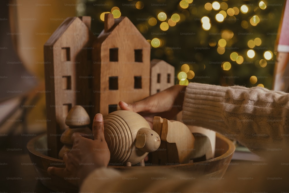 a person holding a wooden toy house in front of a christmas tree