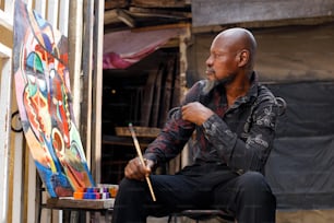 a man sitting in front of a painting holding a paintbrush
