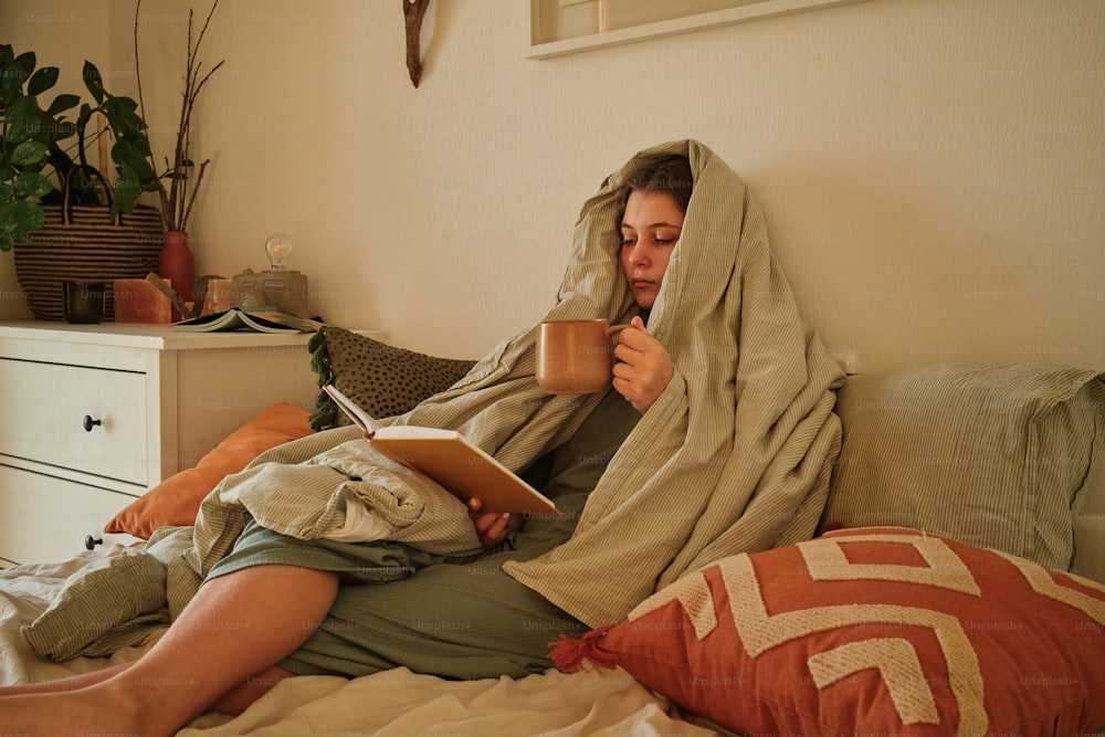 a woman sitting on a bed holding a cup of coffee and reading a book