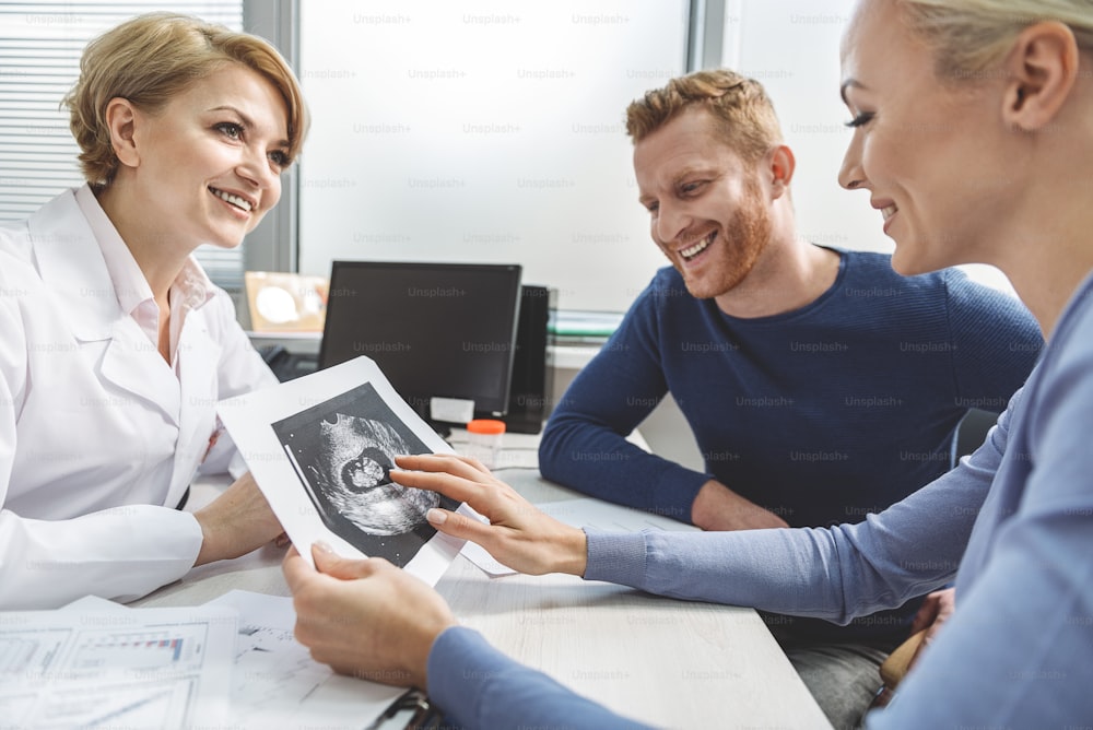 Smiling female gynecologist is sitting afore cheerful clients. Woman holding ultrasound picture and looking with tenderness