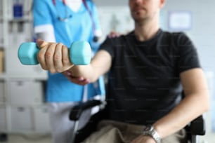 Close-up of physician helping patient to develop joint. Man lifting blue dumbbell sitting in wheelchair in hospital. Modern medicine and physical therapist concept