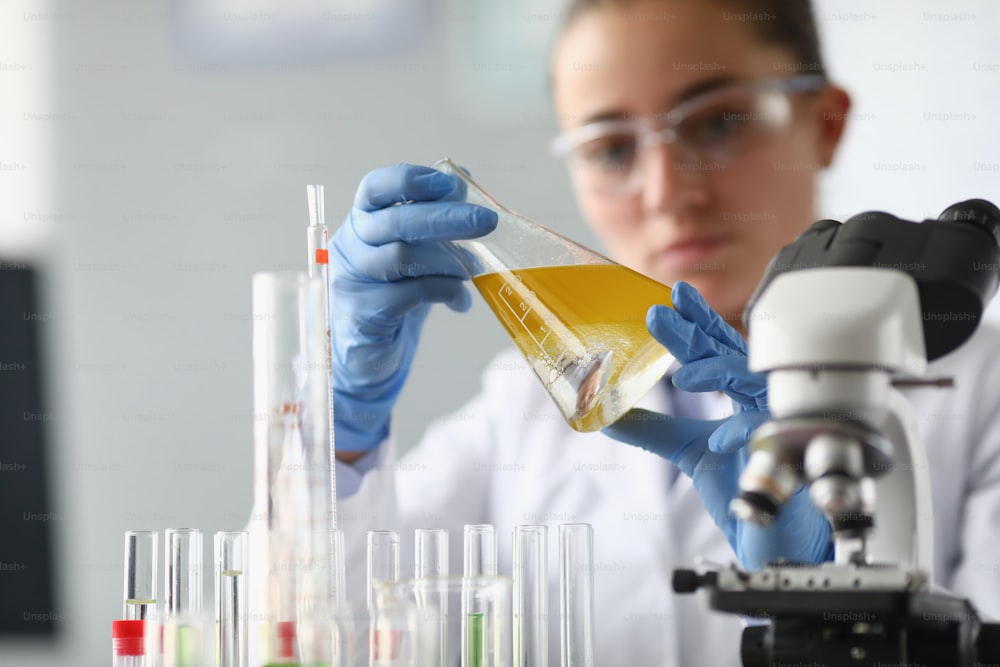 Il chimico della donna tiene il pallone con il liquido giallo nelle sue mani nel laboratorio chimico. Controllo di qualità del concetto di prodotti petrolchimici.