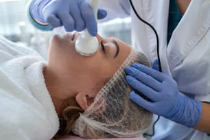 young woman receiving professional procedure facial skin ultrasound cavitation as exfoliation rejuvenation and hydratation from cosmetologist