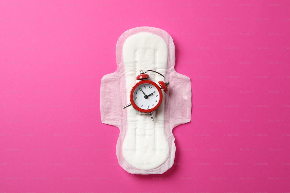 Sanitary pad with alarm clock on pink background, top view