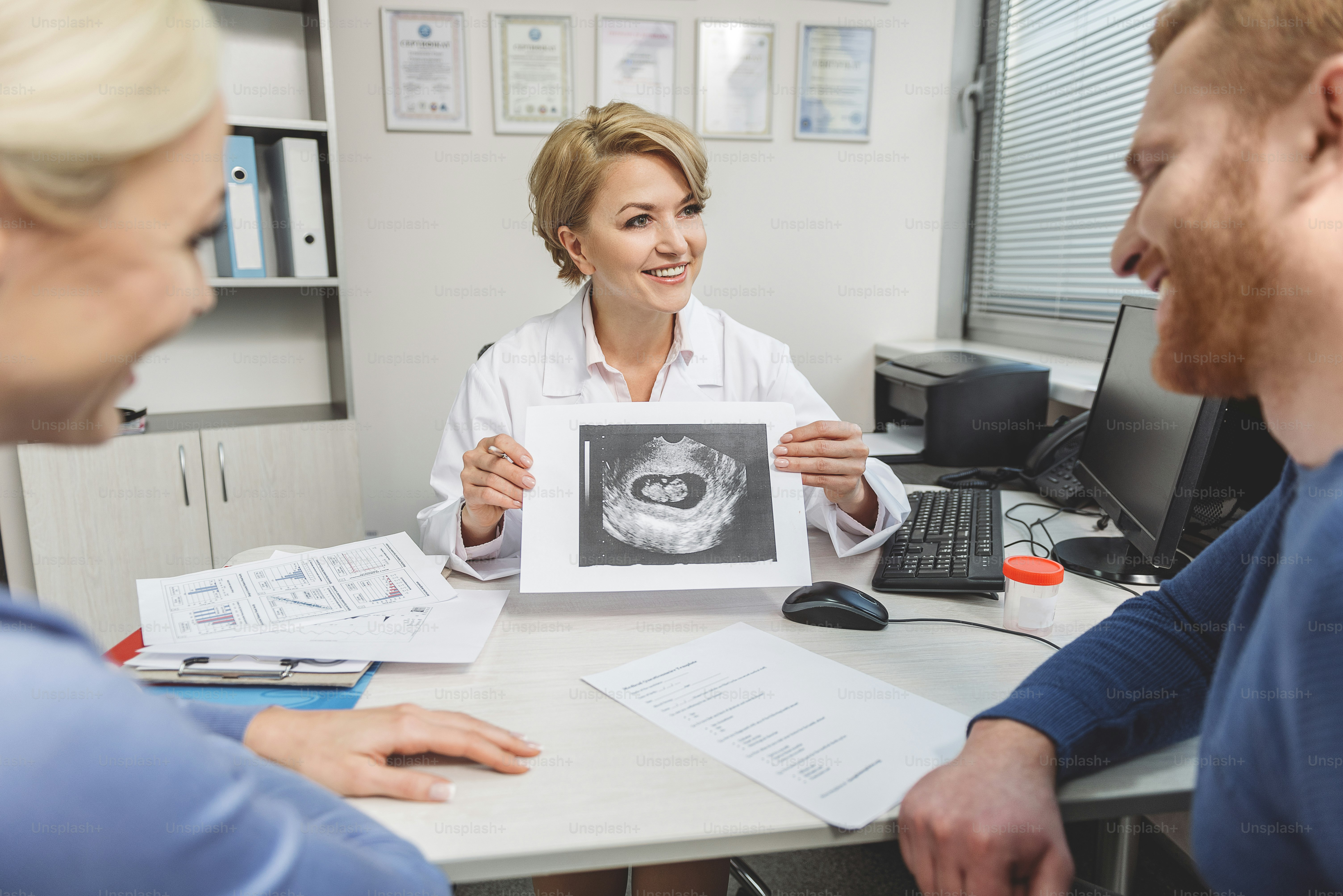 Smiling gynecologist is sitting near table. She holding ultrasound picture and showing it to patients