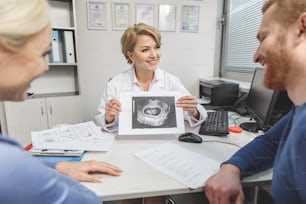 Ginecologista sorridente está sentado perto da mesa. Ela segurando a imagem do ultrassom e mostrando aos pacientes