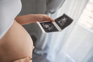Close up shot of big pregnant tummy and woman hand holding sonogram image of healthy unborn baby