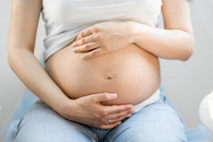 Mujer embarazada abrazando su vientre, sentada en una silla ginecológica durante un examen médico, vista recortada sin rostro enfocado en el abdomen