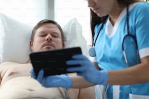 Close-up of concentrated male patient learn about cardiology result. Medical worker in uniform holding modern tablet with heart rate. Hospital bed. Medicine and checkup concept