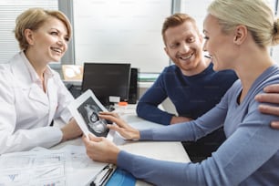 Ein aufmerksamer Arzt sitzt vor der schwangeren Frau und ihrem Partner. Sie hält Ultraschallbild eines Kindes in der Hand