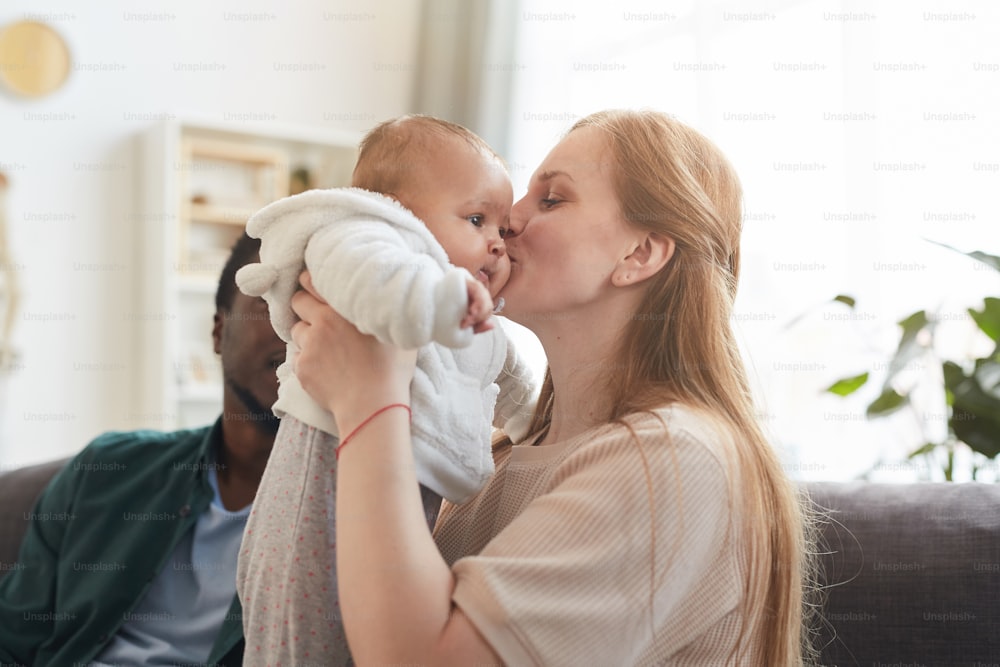 Portrait of happy interracial family at home, focus on Caucasian woman kissing cute baby in foreground, copy space