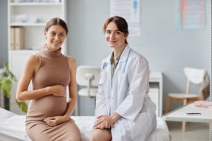 Portrait of pregnant woman and her gynecologist smiling at camera together while sitting on couch at doctors office