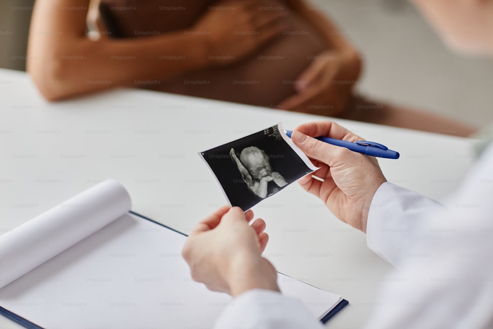 Primo piano del medico che esamina l'immagine dell'ecografia dell'embrione seduto al tavolo e parla con la donna incinta durante la sua visita