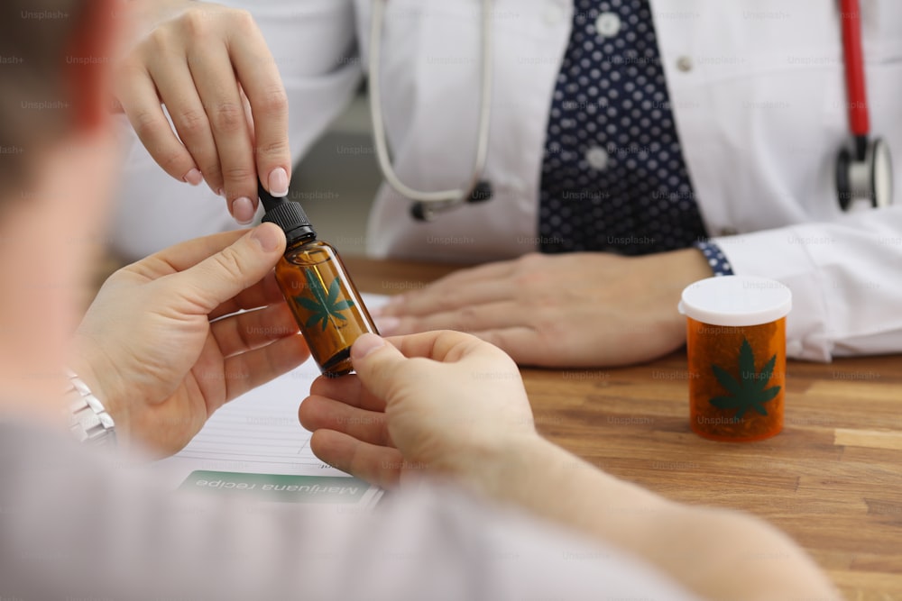 医師の接写は、健康治療のために大麻の抽出油でボトルを与えます。診療所で予約中の男性。代替医療、ヘルスケアの概念