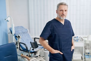 Front view of serious mature doctor posing for camera among gynecological equipment
