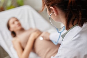 Rear view of female doctor examining the belly of pregnant woman with stethoscope at hospital