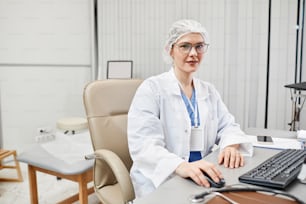 Portrait of female doctor wearing lab coat and looking at camera while sitting at desk in clinic, copy space