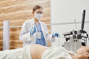 Retrato de una doctora que usa una máquina de ultrasonido y usa una máscara mientras examina a una mujer embarazada en la clínica