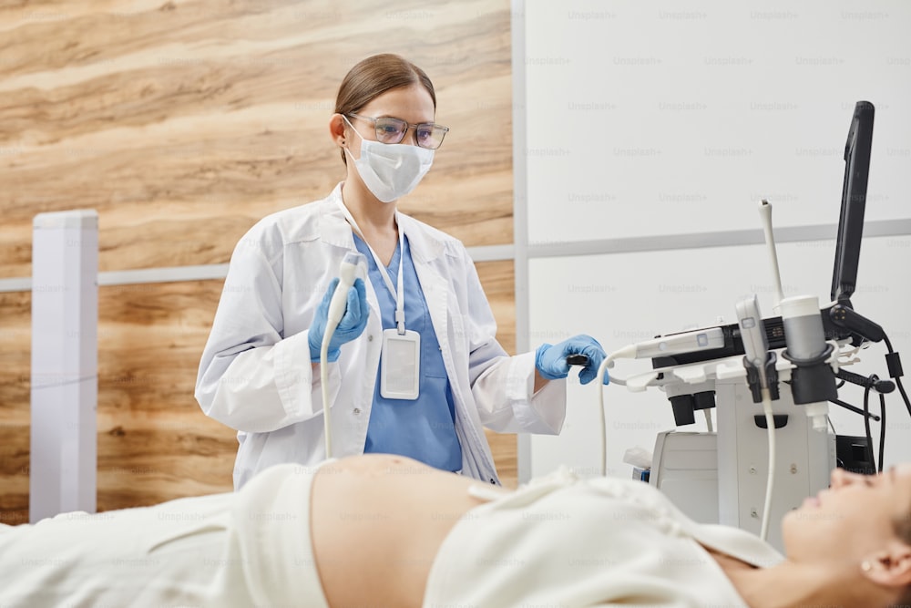 Portrait of female doctor using ultrasound machine and wearing mask while examining pregnant woman in clinic
