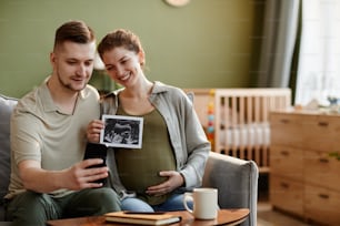 Jeune couple parlant en ligne sur le téléphone portable et partageant leur bonheur avec des proches leur montrant une image échographique