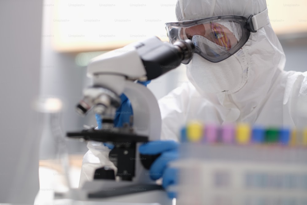 防護服を着た科学者が、近代的な設備の整った実験室で顕微鏡を覗いています。科学研究のための高度な技術でウイルスの進化を研究する研究室助手