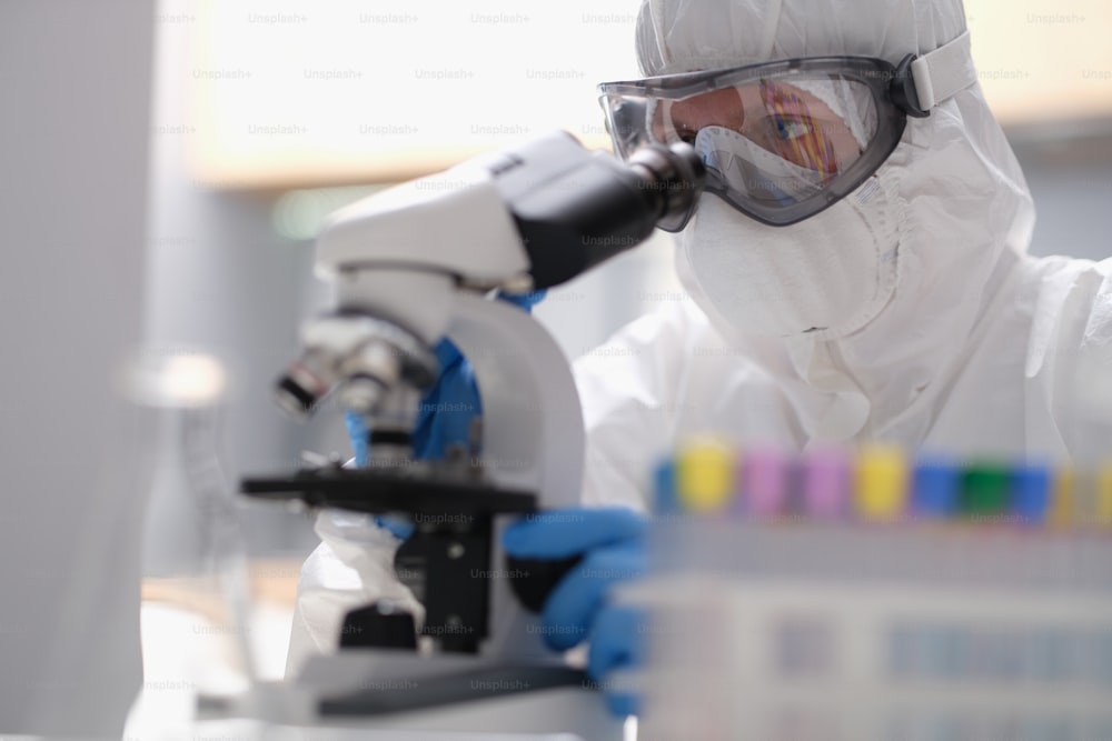 Scientist in a protective suit looking through a microscope in modern equipped laboratory. Laboratory assistant studying the evolution of the virus using high technology for scientific research