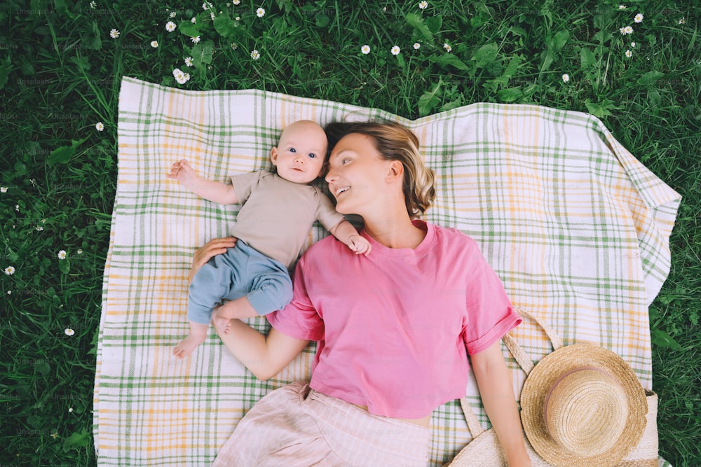 Smiling mom and baby lying on blanket on green grass at summer. Family relaxing and having picnic outdoors. Beautiful mother with her baby on nature. Concept of motherhood, human happiness, eco life.