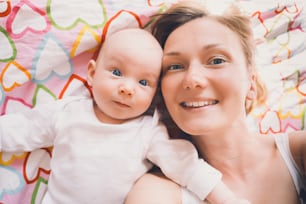 Portrait of mother and baby, top view. Smiling mom takes selfie of herself and her cute little baby on blanket with pattern of hearts. Young woman enjoying motherhood.