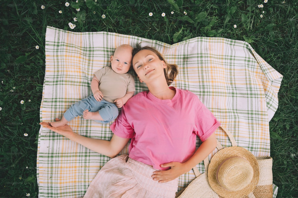 Smiling mom and baby lying on blanket on green grass at summer. Family relaxing and having picnic outdoors. Beautiful mother with her baby on nature. Concept of motherhood, human happiness, eco life.