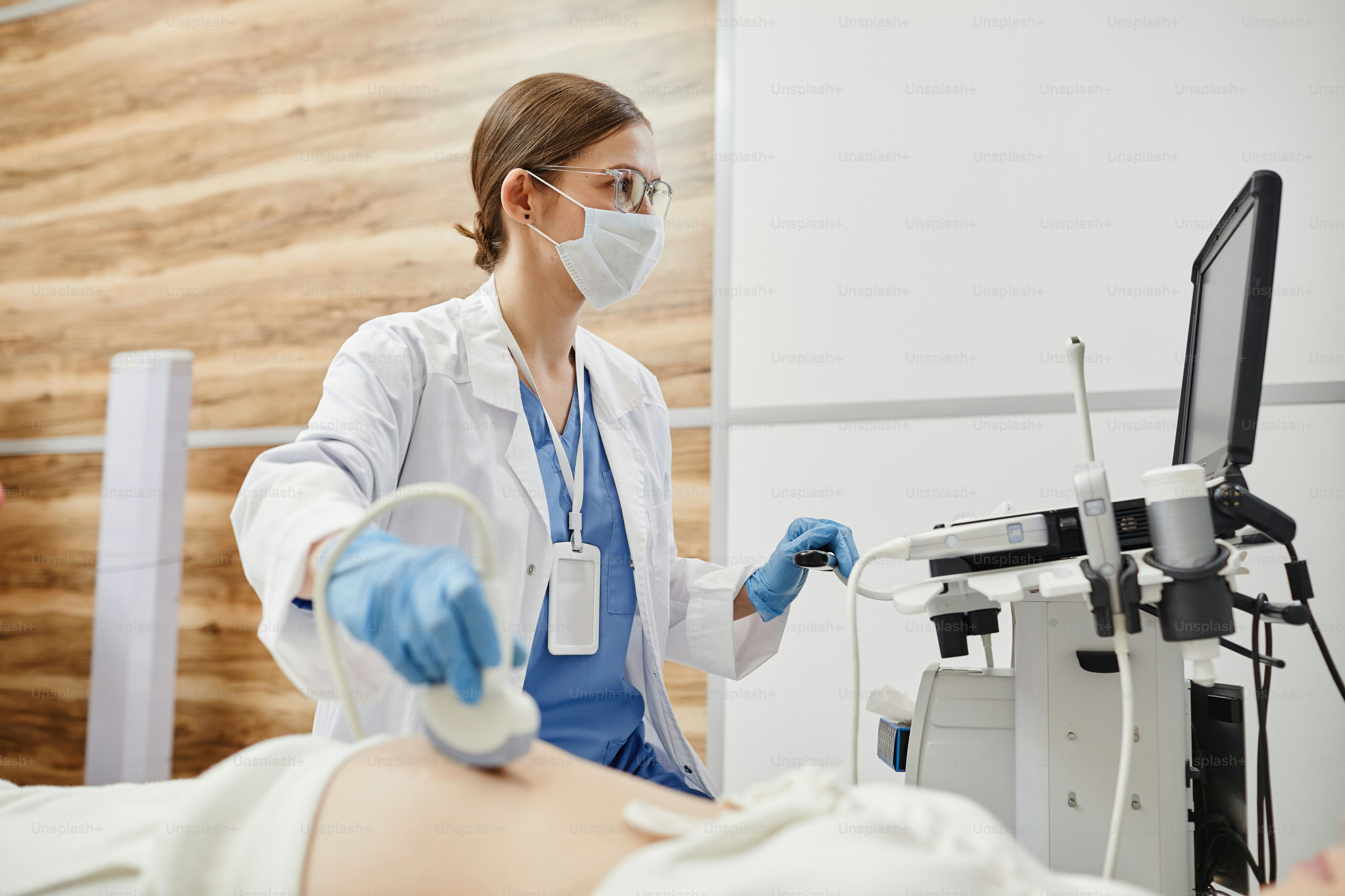 Side view portrait of female doctor using ultrasound machine and wearing mask while examining pregnant woman in clinic