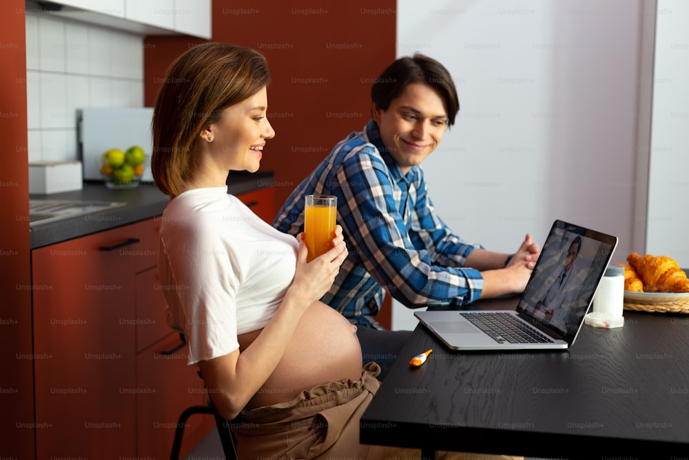Smiling man and pregnant woman are sitting in kitchen and enjoying online conference with doctor
