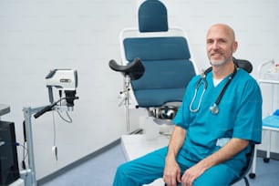 Smiling gynecologist is sitting in a medical office while he is wearing a uniform and a stethoscope is hanging around his neck