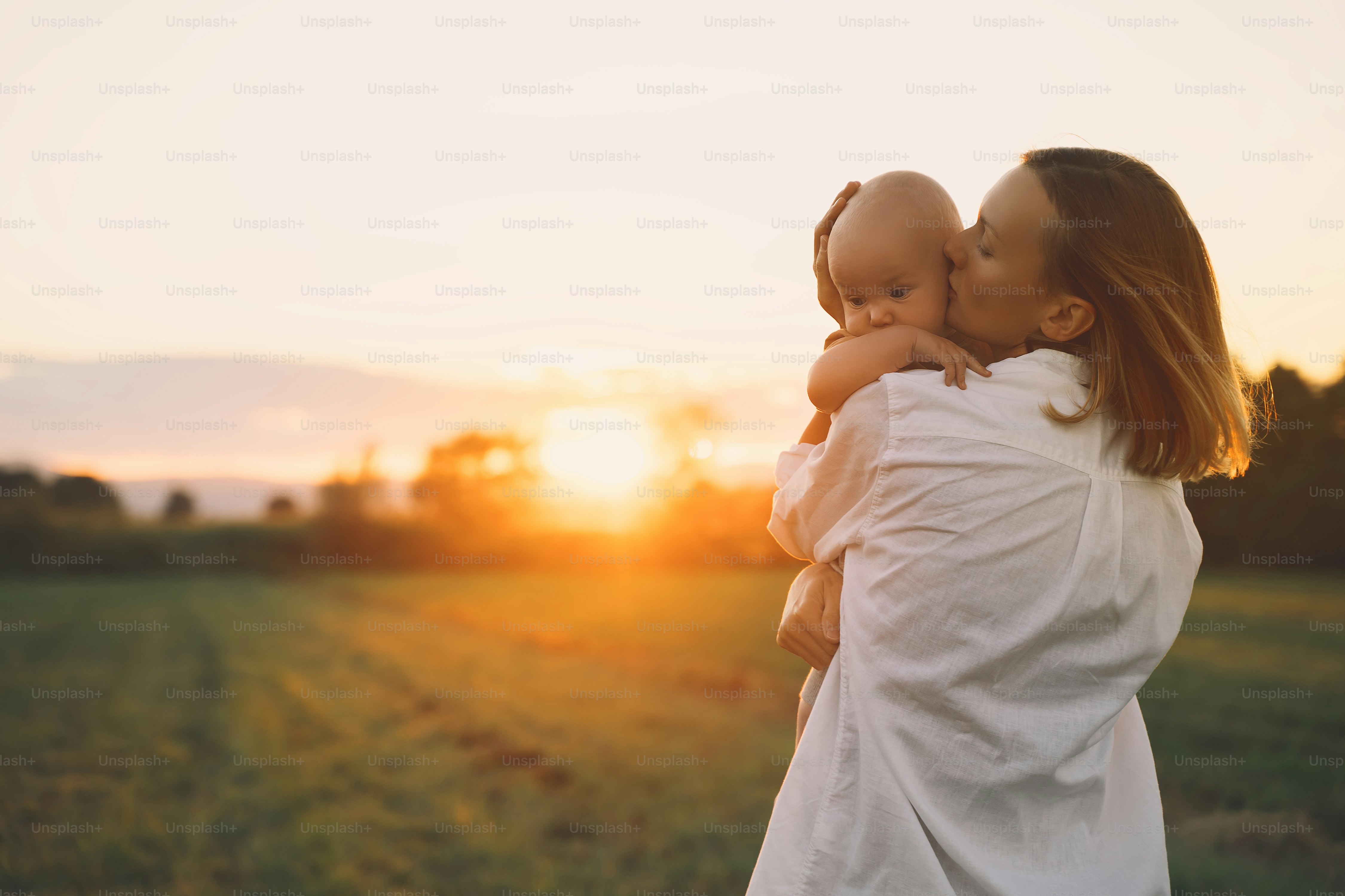 Ibu dan bayi yang penuh kasih saat matahari terbenam. Wanita cantik dan anak kecil dengan latar belakang alam. Konsep keibuan alami. Keluarga sehat yang bahagia di musim panas di luar ruangan. Emosi dan perasaan manusia yang positif.