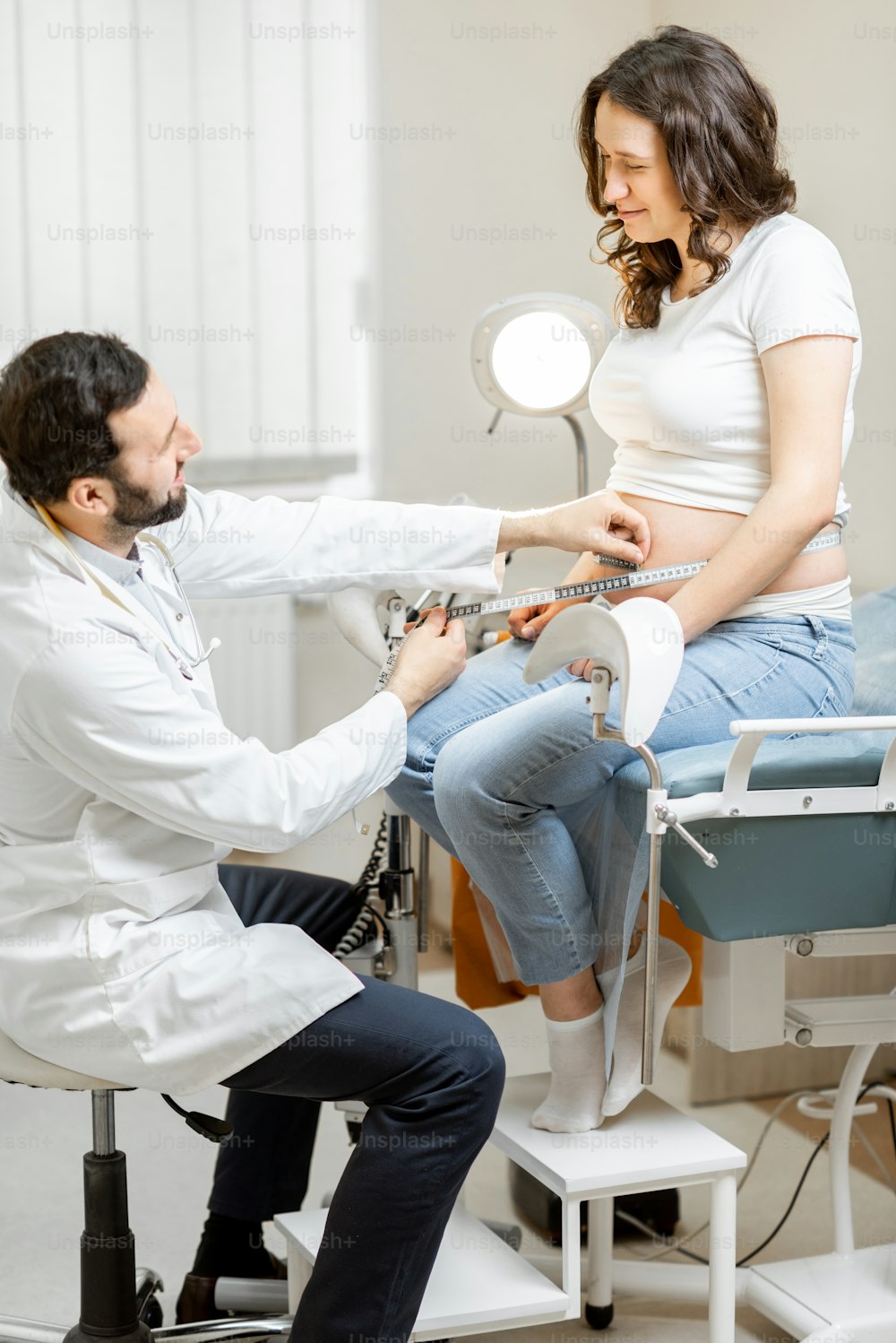 Male doctor measuring pregnant woman's belly with a tape during a medical examination in the office. Concept of medical care and health during a pregnancy